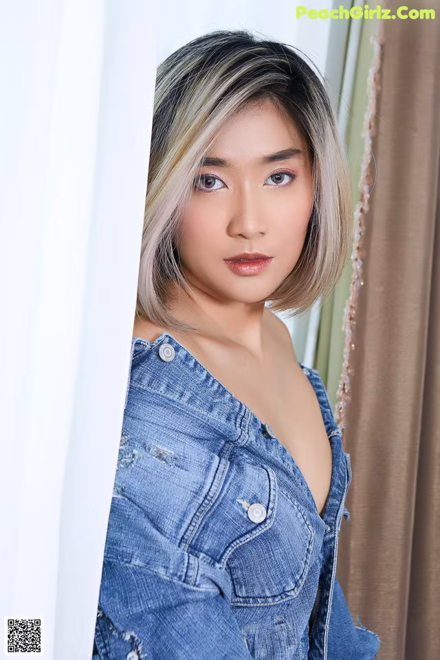 A woman in a denim jacket leaning against a curtain.